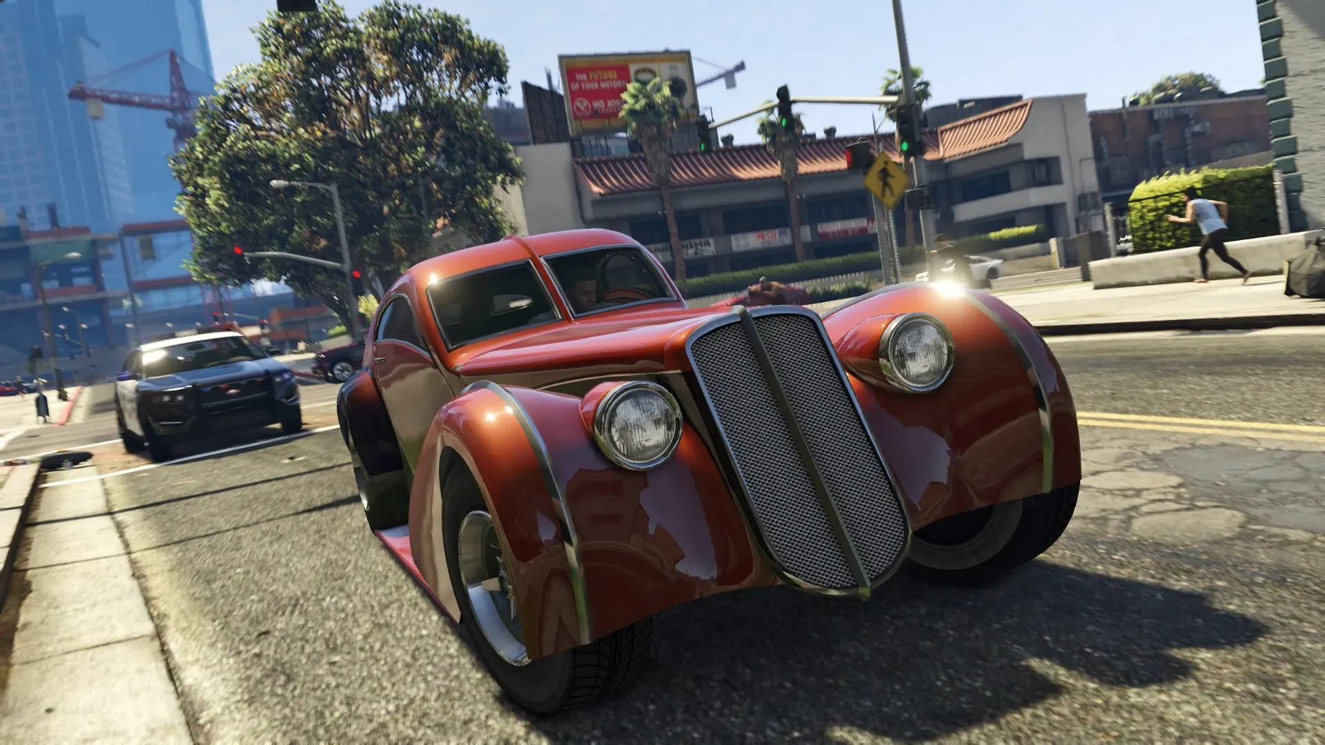 A red 1930s style coupe swerves through a city intersection with a police car close behind