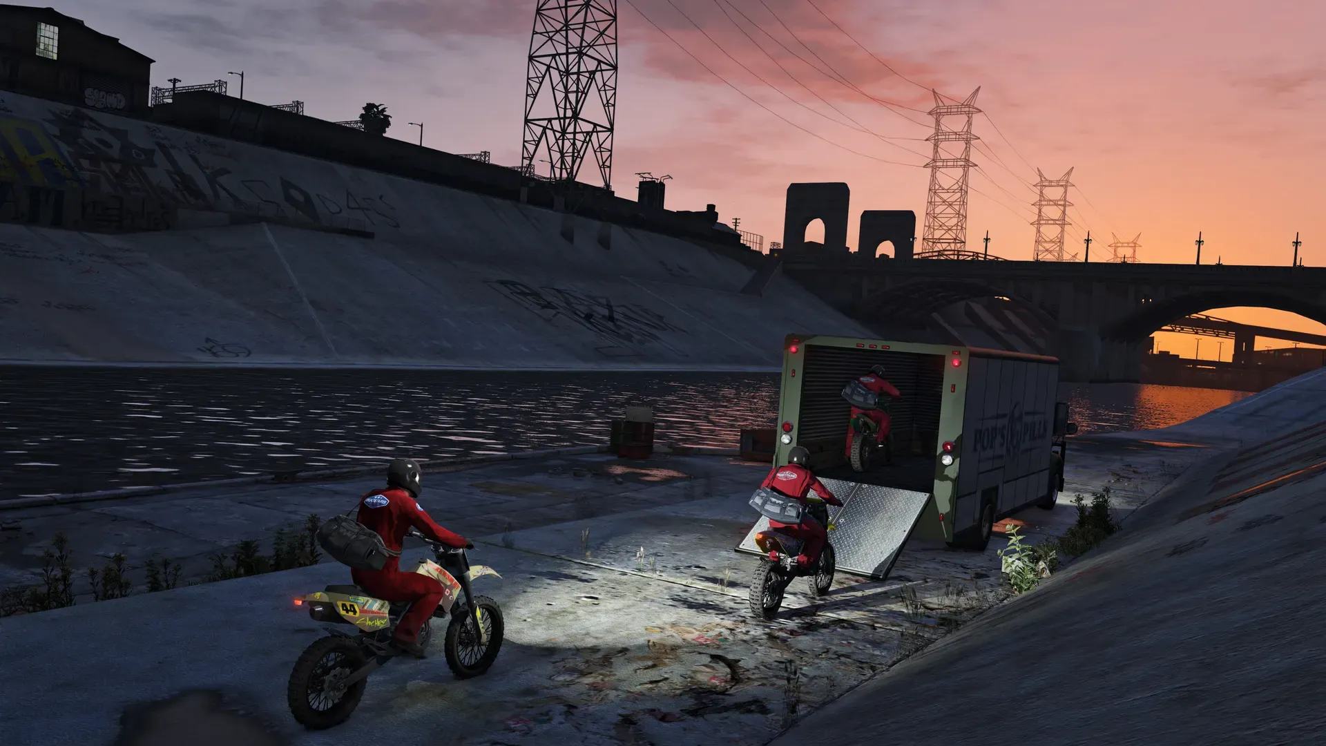 Two dirt bike riders race out of a truck along the concrete Los Santos river channel at sunset