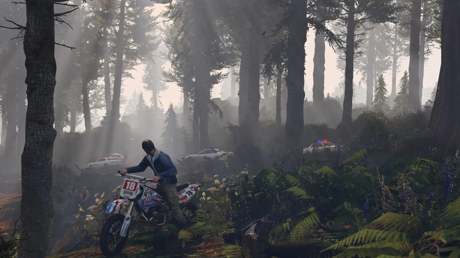 A rider pushes a dirt bike through a misty pine forest while police cars wait on the road behind