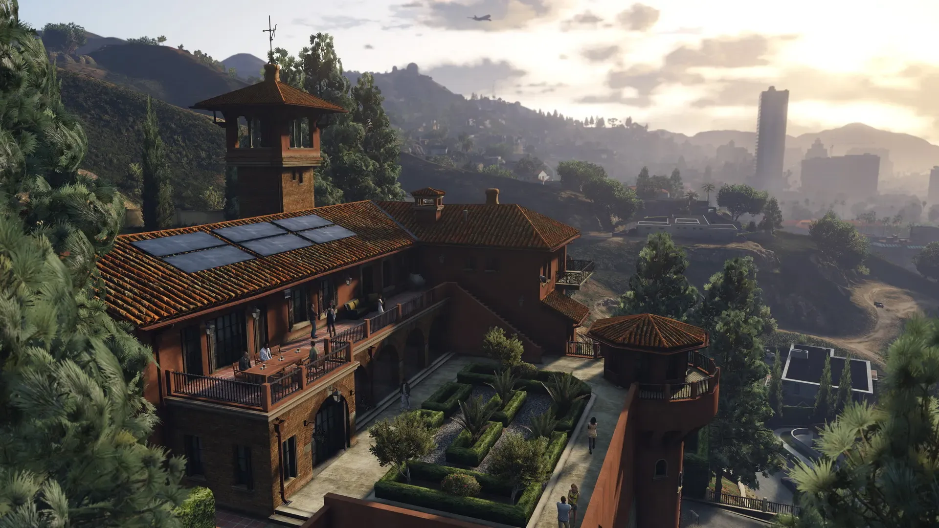 A large terracotta roofed mansion with a tower and formal gardens on a wooded hillside