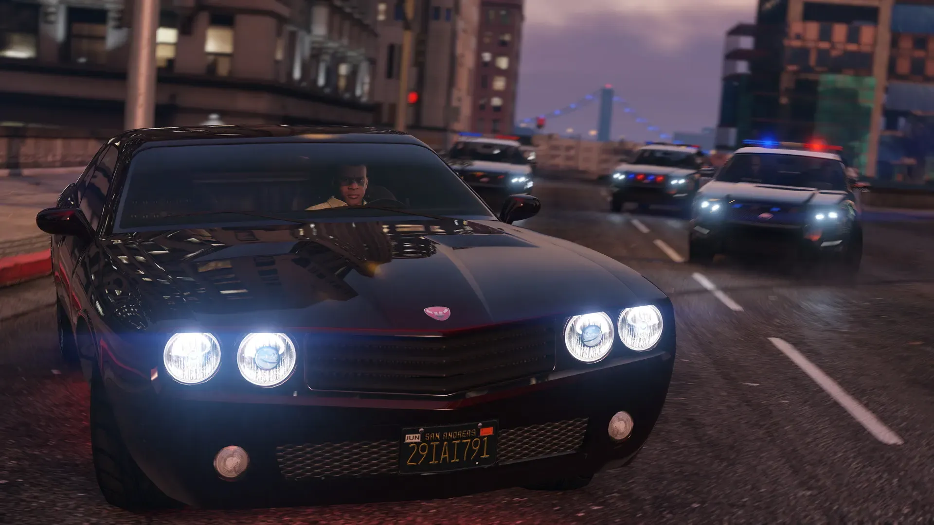 A black muscle car with quad headlights speeds ahead of several police cruisers on a city street at dusk