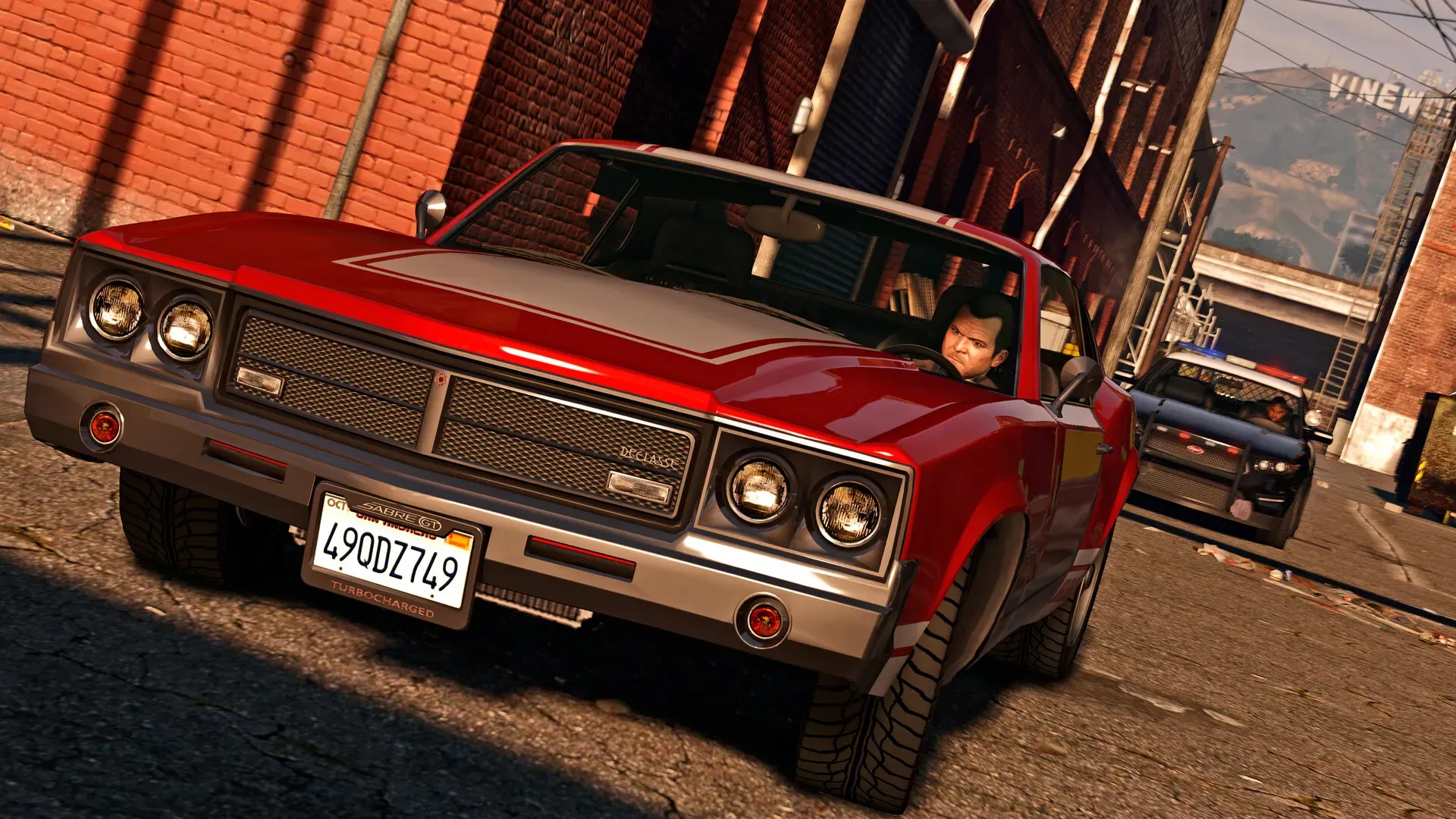 A driver in sunglasses steers a red classic convertible down an alley as a police car follows, with the Vinewood sign in the distance