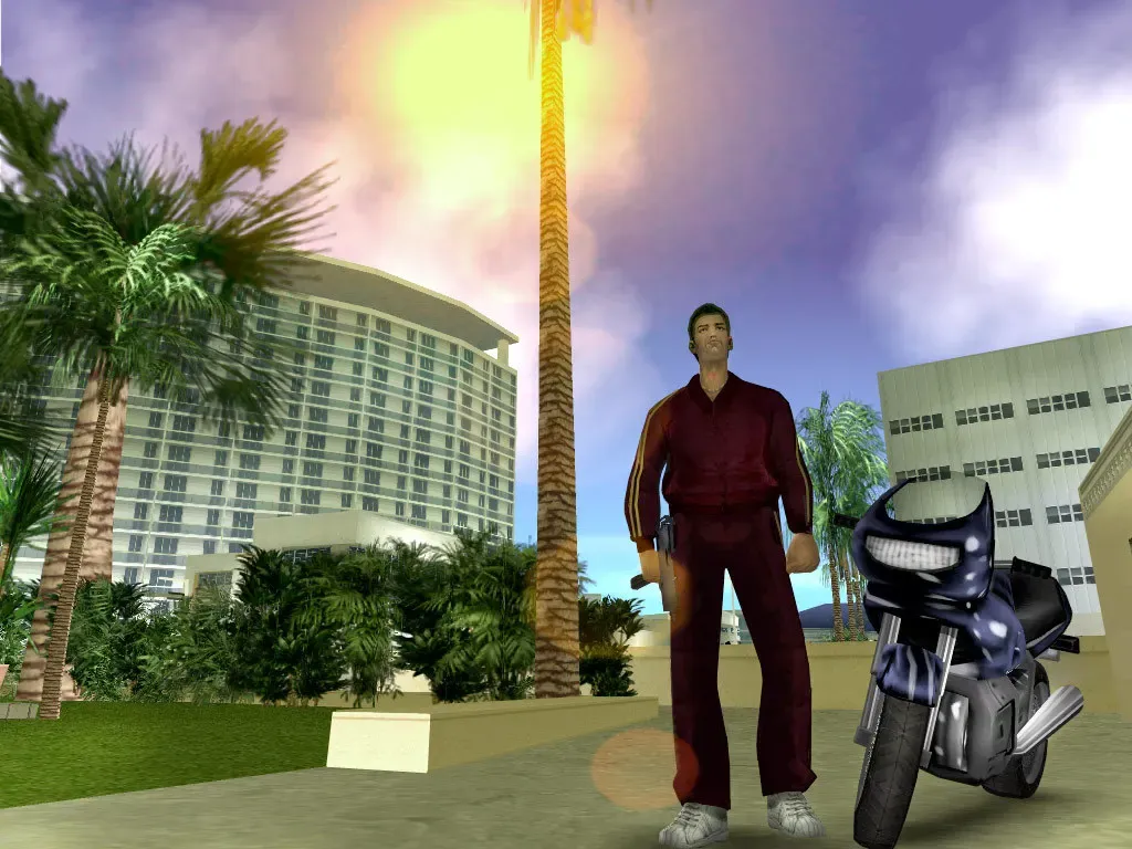 Tommy in a dark red tracksuit stands beside a dark blue sports motorbike under palm trees, with a curved white hotel behind him