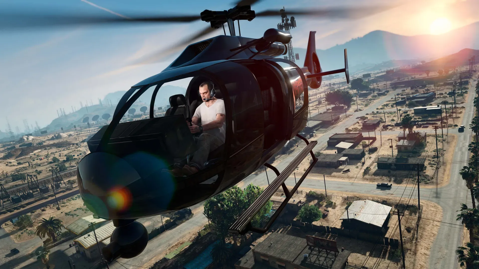 Trevor pilots a red and black helicopter low over a dusty desert town at sunset