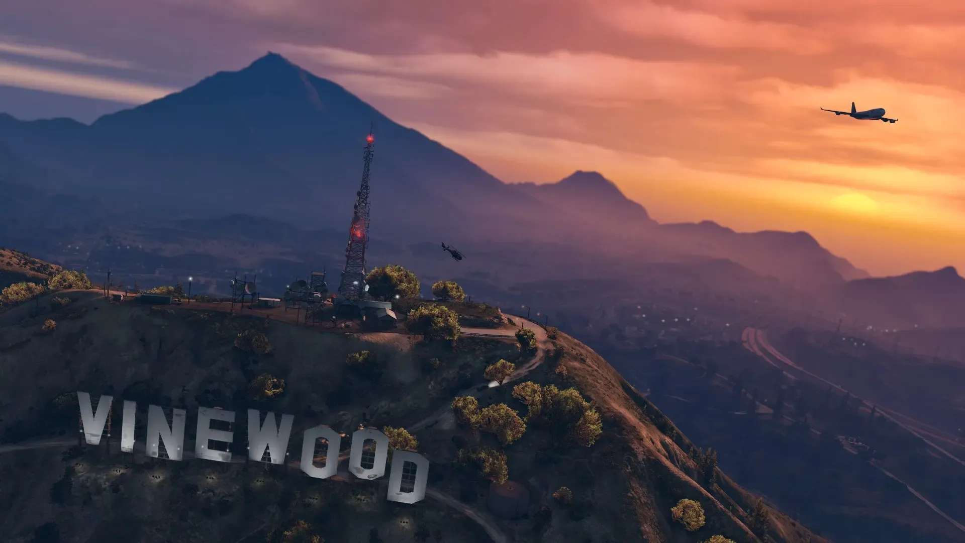 The Vinewood sign on its hilltop below a radio mast at sunset, with a jet crossing an orange sky