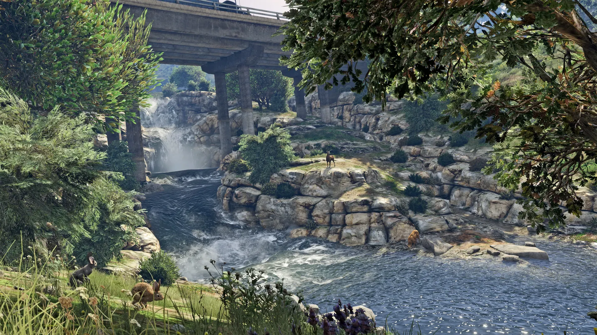 A waterfall spills into a rocky stream beneath a road bridge, with deer drinking among the trees